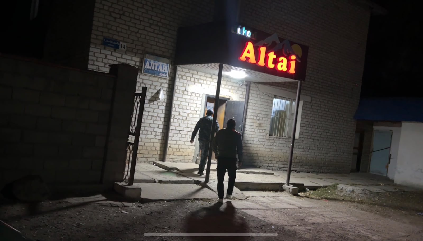 Двое мужчин заходят в здание с неоновой вывеской «Altai» в ночное время.