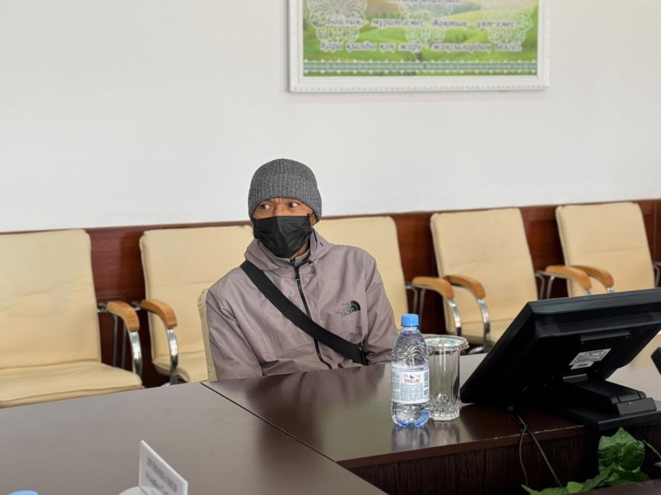 Күршілі бөлмеде үстел басында отырған, бетперде мен сұр бас киім киген ер адам.