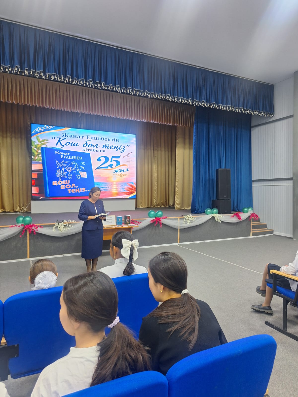 Сахнада Жанат Елшібектің кітабының таныстырылымы өтіп жатыр, ал алдында оқушылар отыр.
