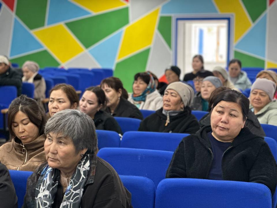 Көк орындықтарда отырған әйелдер тобы. Фонда түрлі-түсті геометриялық оюлы қабырға бейнеленген.
