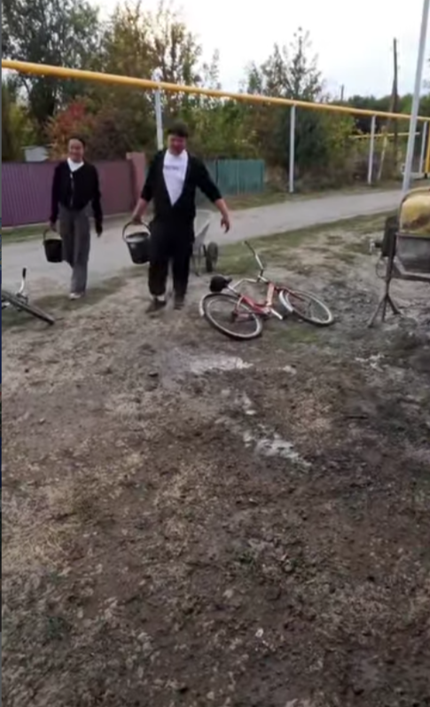 Қолына шелек ұстаған, костюм киген екі ер адам және жерде жатқан велосипед.