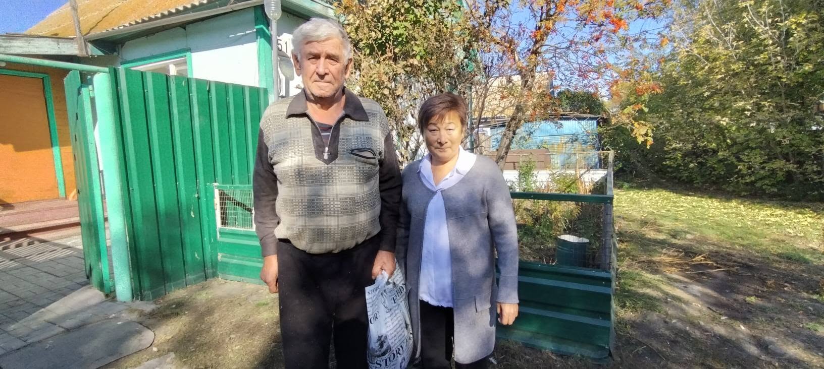 Үйдің алдында тұрған қарт ер адам мен әйелдің суреті.