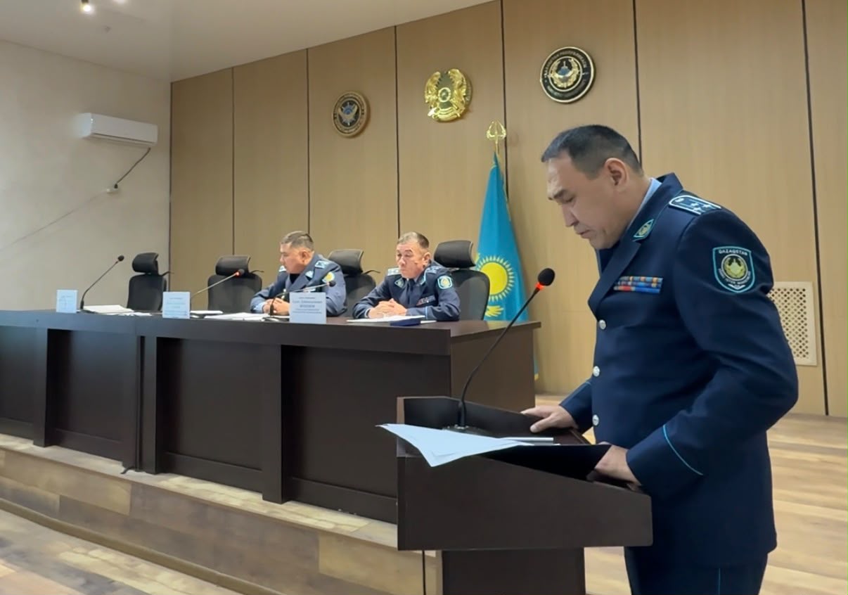 Форма киген офицер трибунада баяндама жасап тұр, ал артында екі әріптесі отыр.