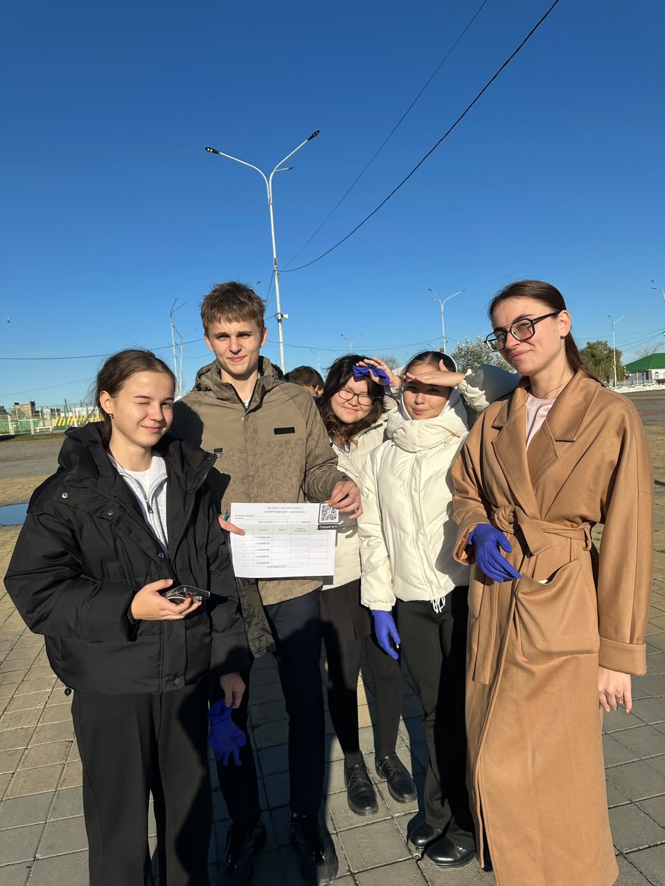 Күн шуағы астында тұрған, қолына құжат ұстаған жастар тобы.