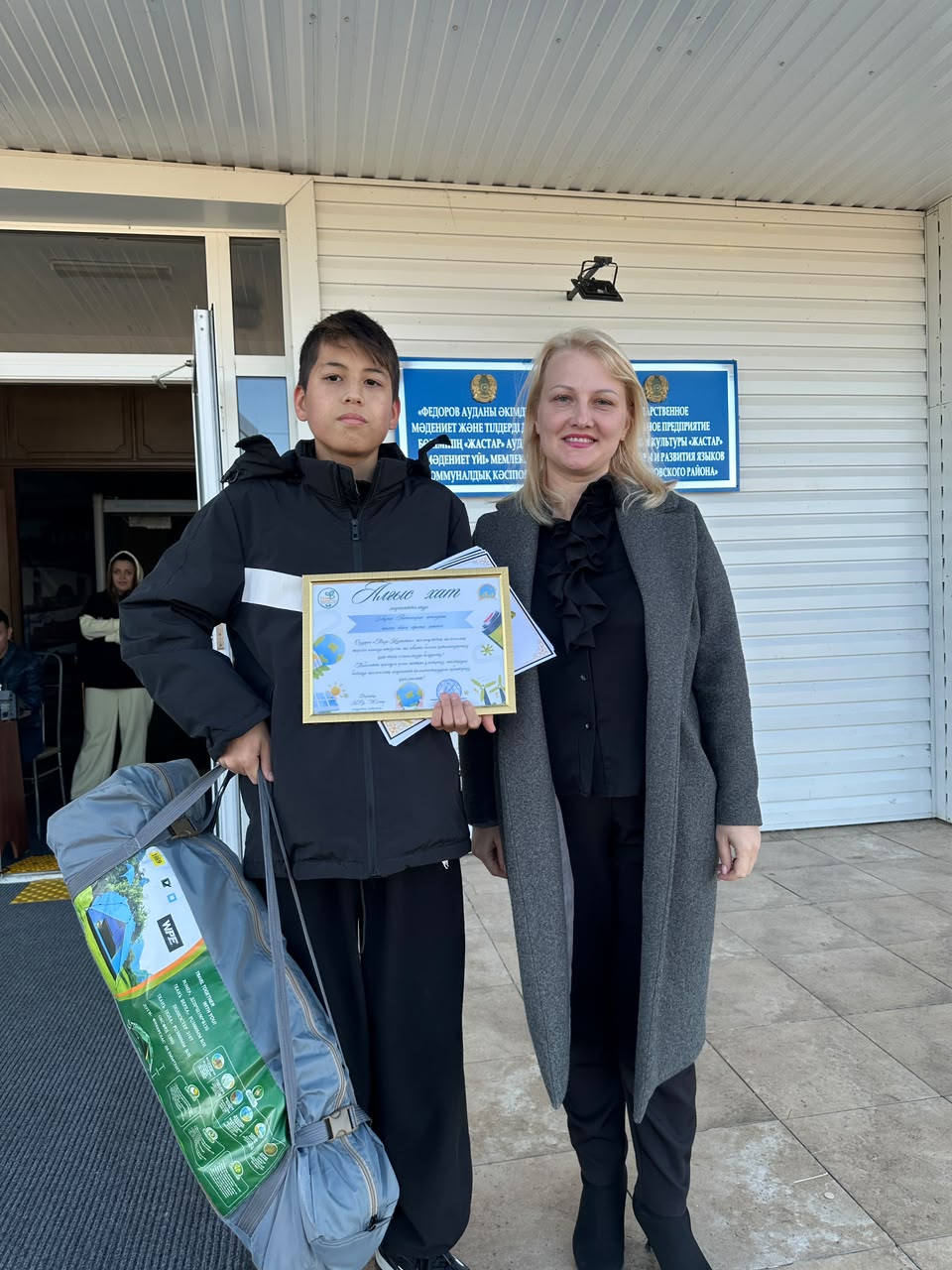 Құттықтау грамотасын ұстаған оқушы мен әйел адамның суреті.
