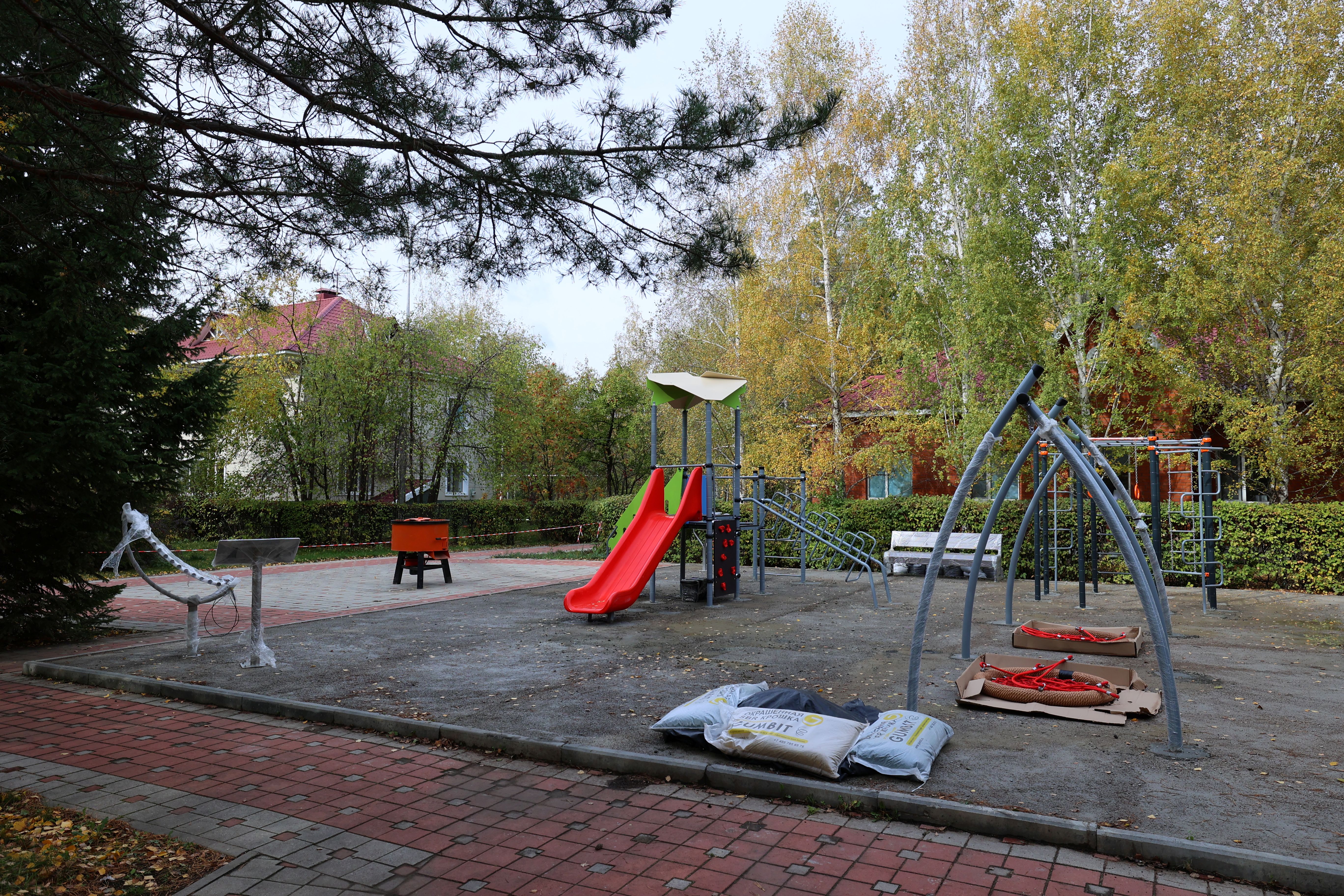Күзгі саябақтағы балалар ойын алаңы: қызыл сырғана, спорттық құрылғылар және құрылыс материалдары.