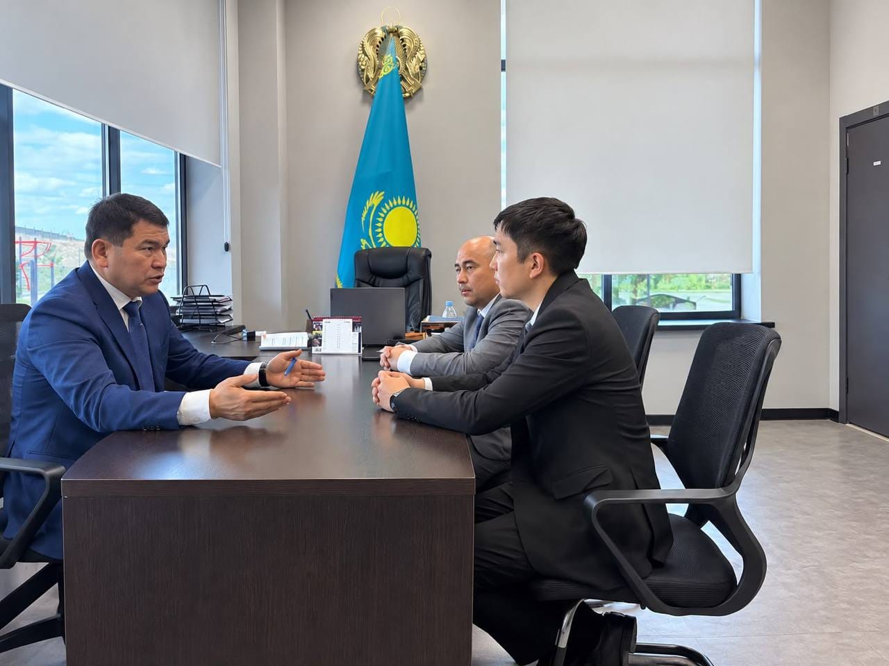 Қазақстан туы тұрған кеңседе костюм киген үш ер адамның жұмыс үстеліндегі кездесуі.