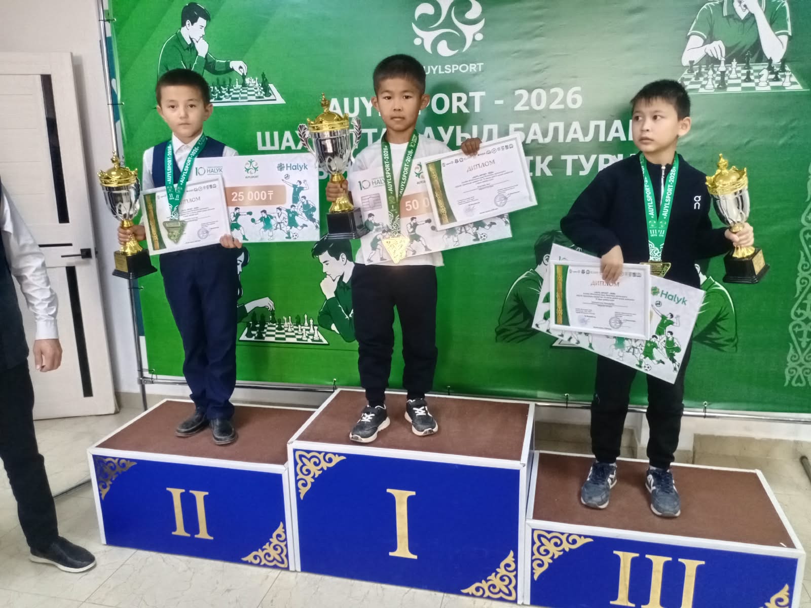 Шахмат турнирінің жүлдегерлері: жеңімпаз үш бала кубок пен диплом ұстап, пьедесталда тұр.