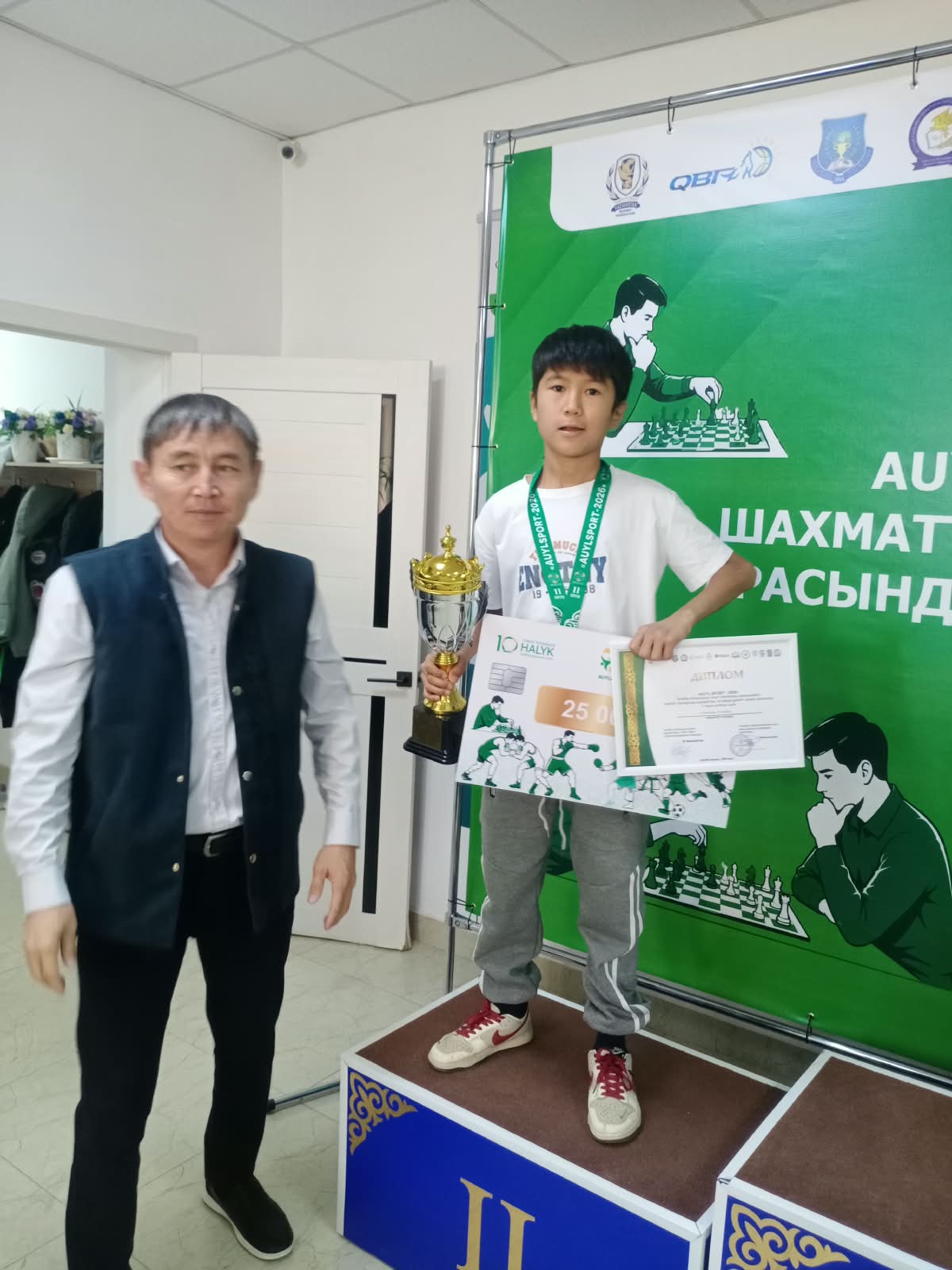Шахмат турнирінің жеңімпазы кубок пен диплом ұстап, пьедесталда тұр, жанында ерекелеген ер адам тұр.