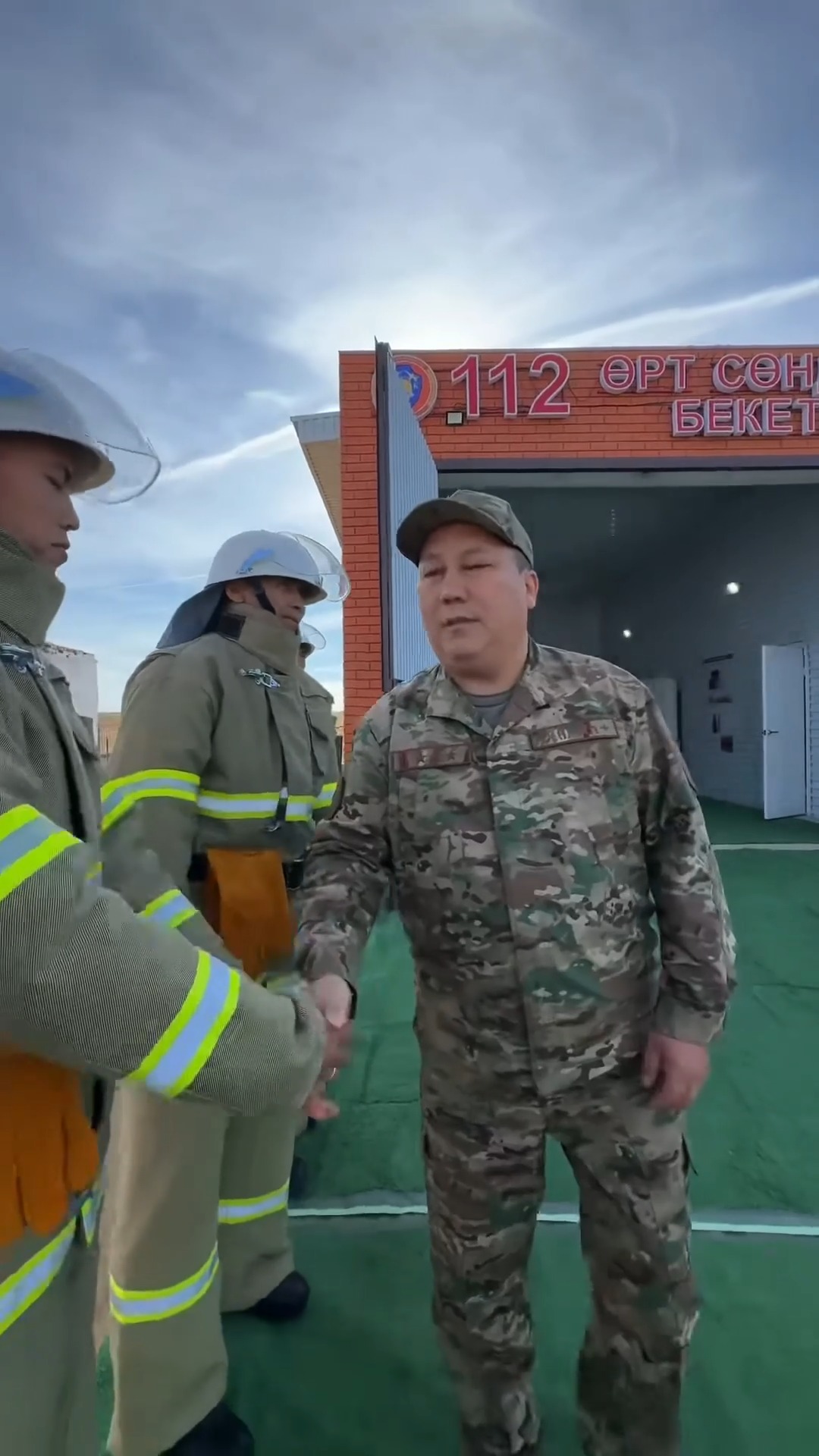 Әскери формадағы ер адамның өрт сөндірушілермен амандасуы. Фонда «112 Өрт сөндіру бекеті» жазуы бар ғимарат.