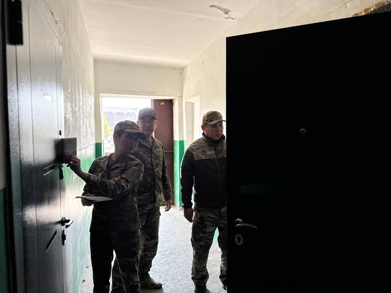 Әскери формадағы үш адам ғимарат дәлізінде тұр, біреуі есікті ашып жатыр.