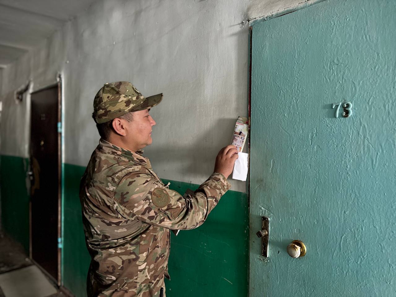 Әскери формадағы адамның үй есігіне хабарландыру қағазын іліп жатқан сәті.