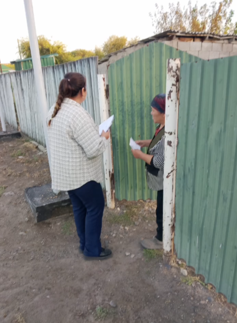 Жасыл қоршаудың жанында қолдарында қағаздары бар екі әйел сөйлесіп тұр.