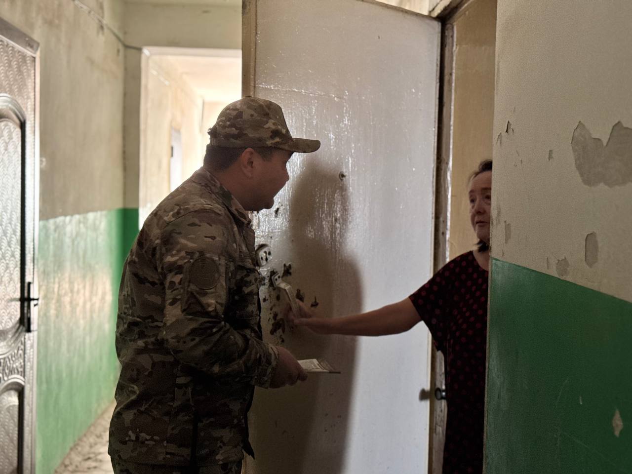 Әскери формадағы адам есік алдында тұрған әйелге құжат немесе қағаз табыстап жатыр.