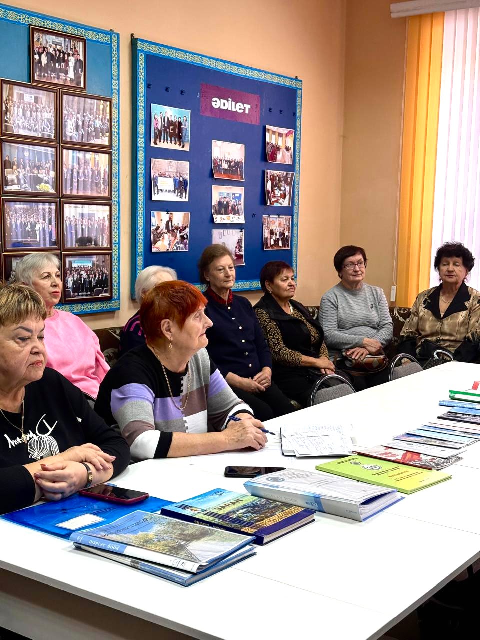 Бір топ әйелдер үстелде қабырғаларында фотосуреттері бар бөлмеде құжаттармен отырады.