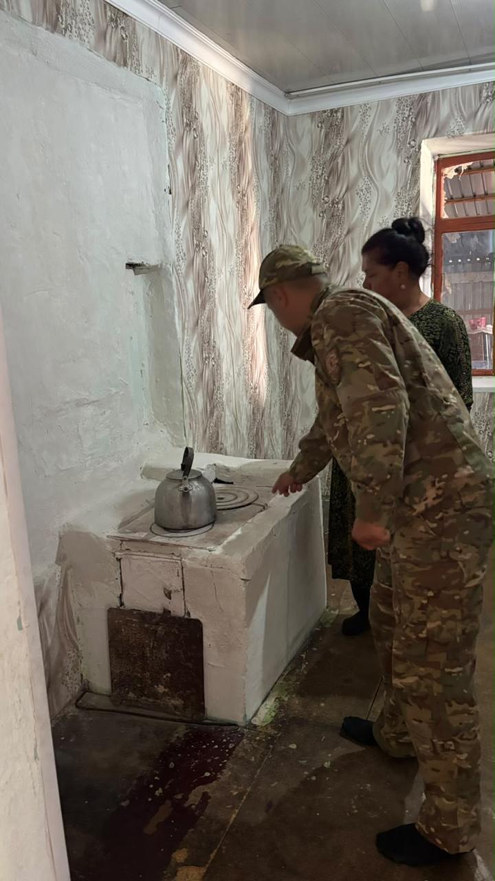Военный в камуфляже и женщина стоят у старой белой печи с чайником в комнате.