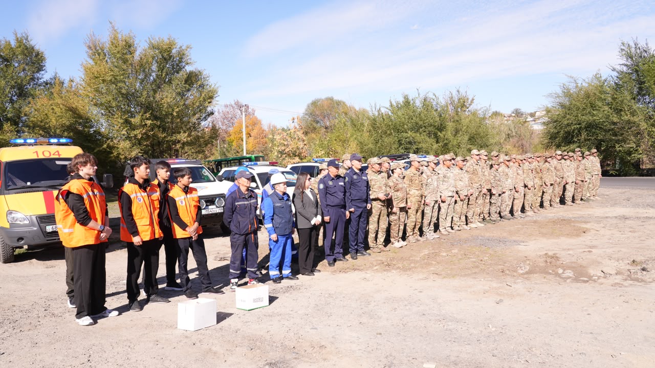 Группа спасателей в оранжевых и синих жилетах и строй военных в камуфляже стоят на открытой площадке рядом со спецтехникой.