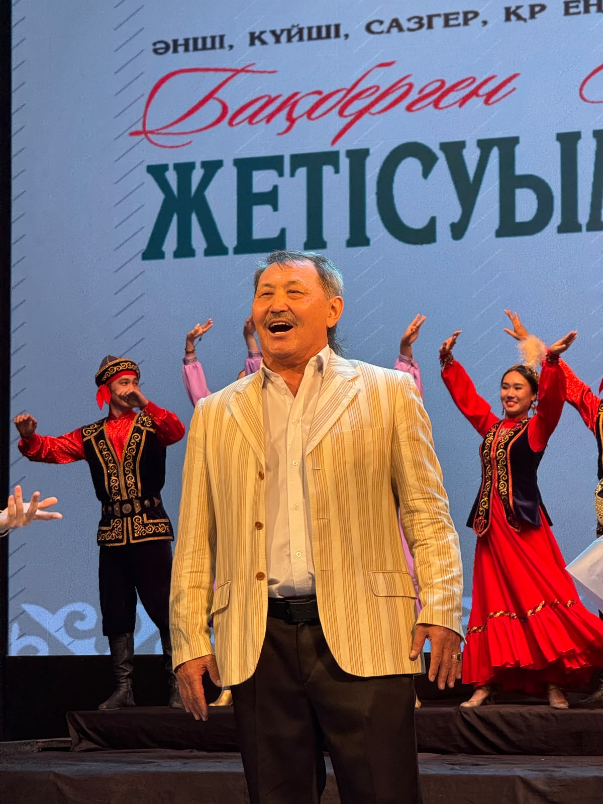 Сахнада ұлттық киімдегі бишілердің алдында тұрған күлімсіреген ер адам.
