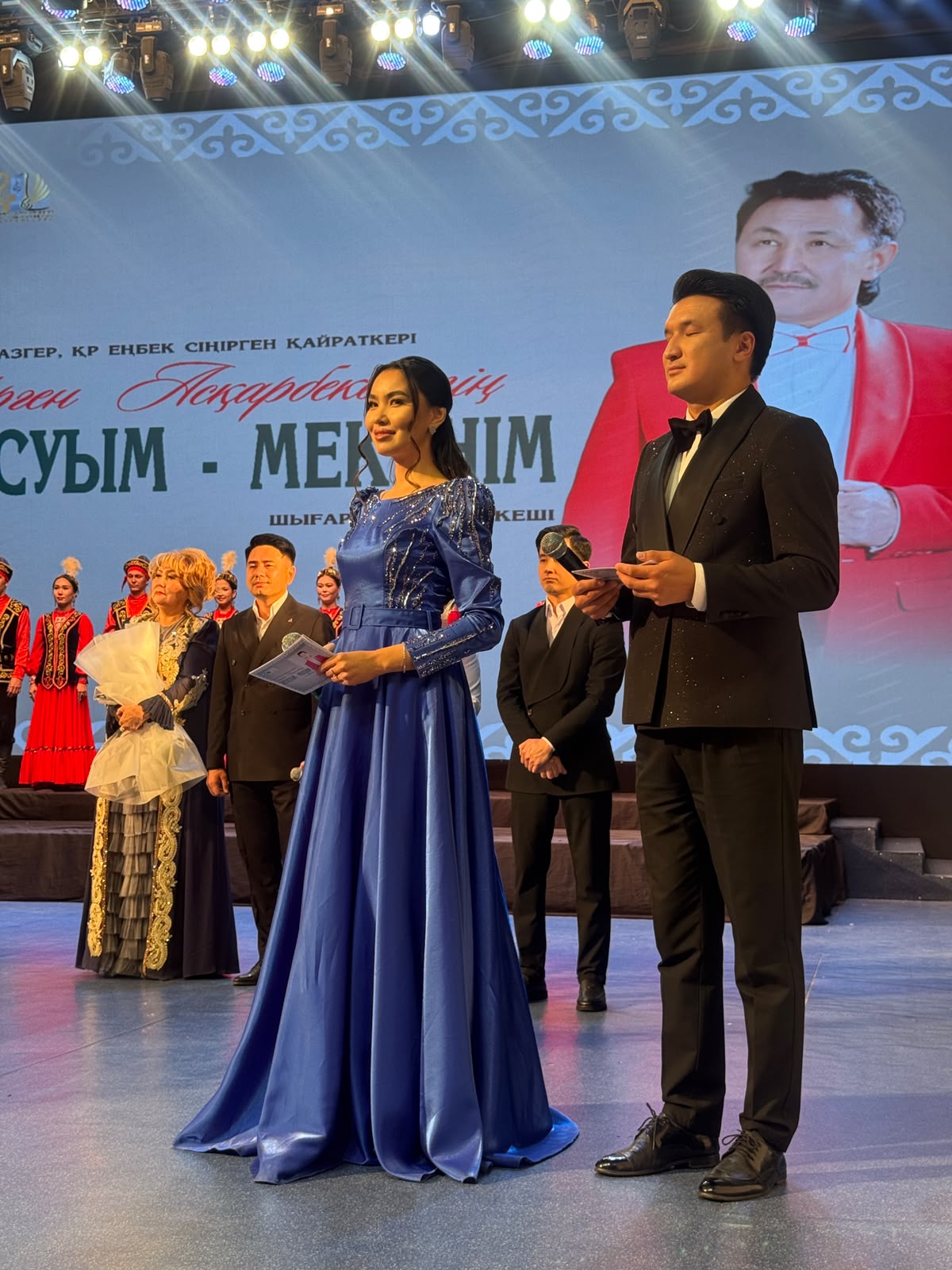 Көк көйлек киген әйел мен смокинг киген ер адам сахнада тұр.