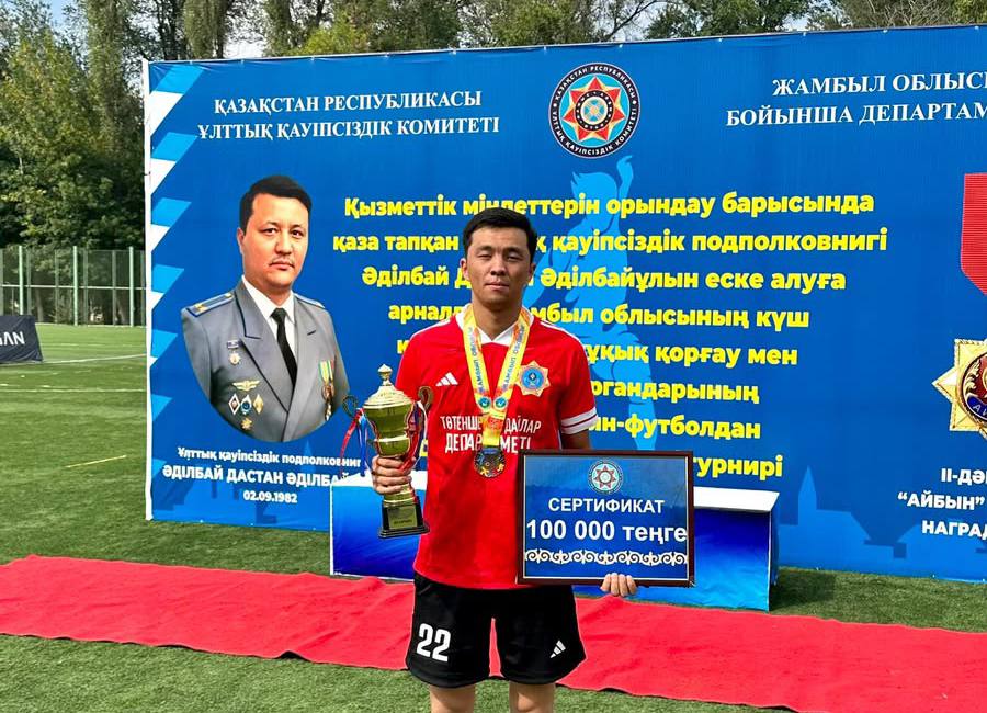 Футбол турнирінің жеңімпазы кубок пен сертификат ұстап тұр. Фонда ҰҚК баннері мен фотосурет бейнеленген.