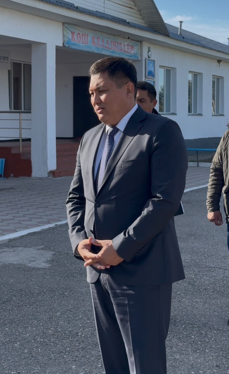 Көк жиектегі костюм киген ер адамның ғимарат алдында тұрған суреті.