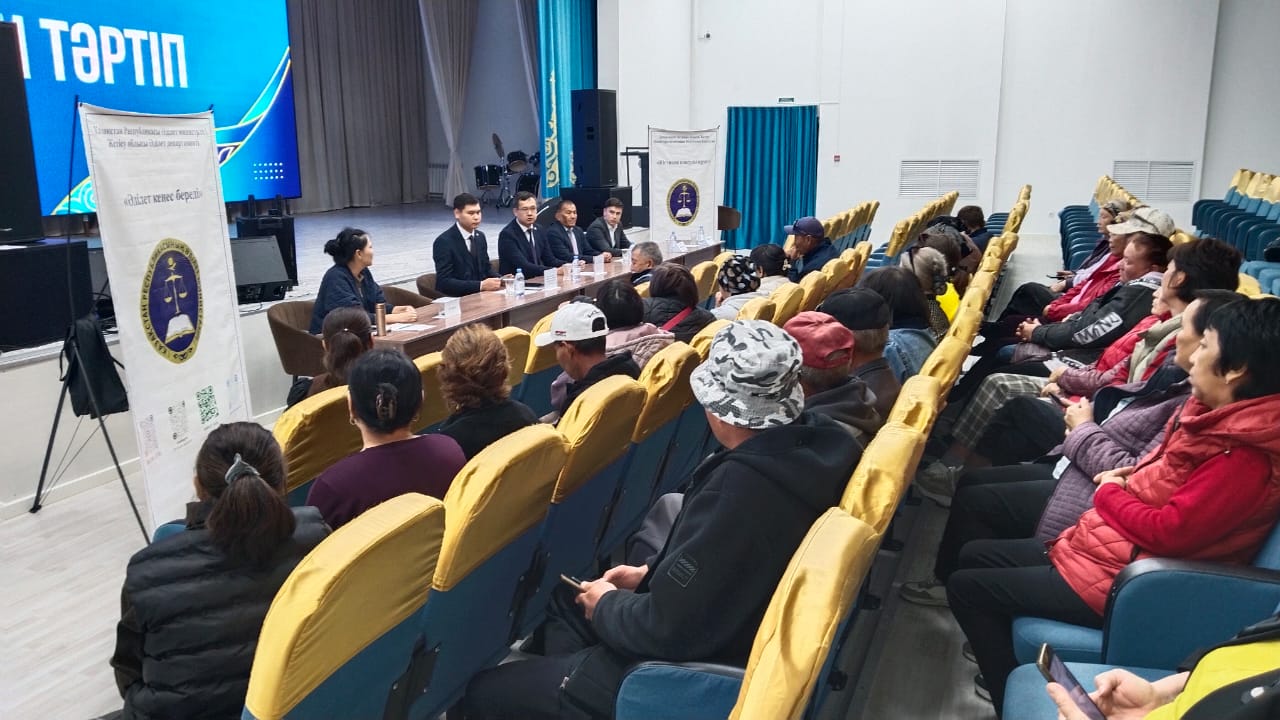 Люди на конференции в зале: спикеры за столом перед экраном и слушатели в желтых креслах.