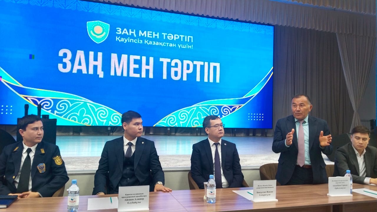 Пять мужчин в костюмах на пресс-конференции на фоне синего экрана с надписью «ЗАҢ МЕН ТӘРТІП».