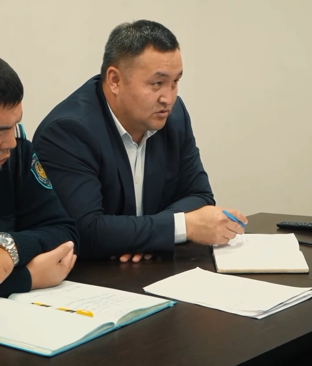 Көк костюм киген ер адам үстел басында отырып, дәптерге жазып жатыр.