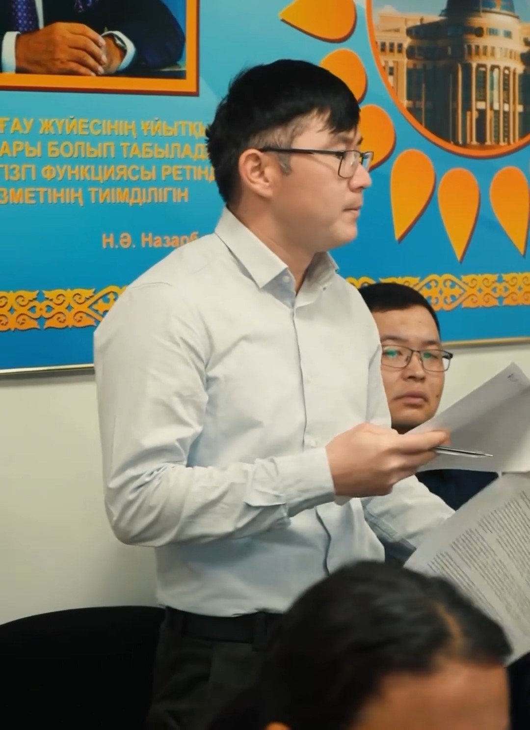Қолына қағаз ұстап, сөйлеп тұрған көзілдірікті ер адам.