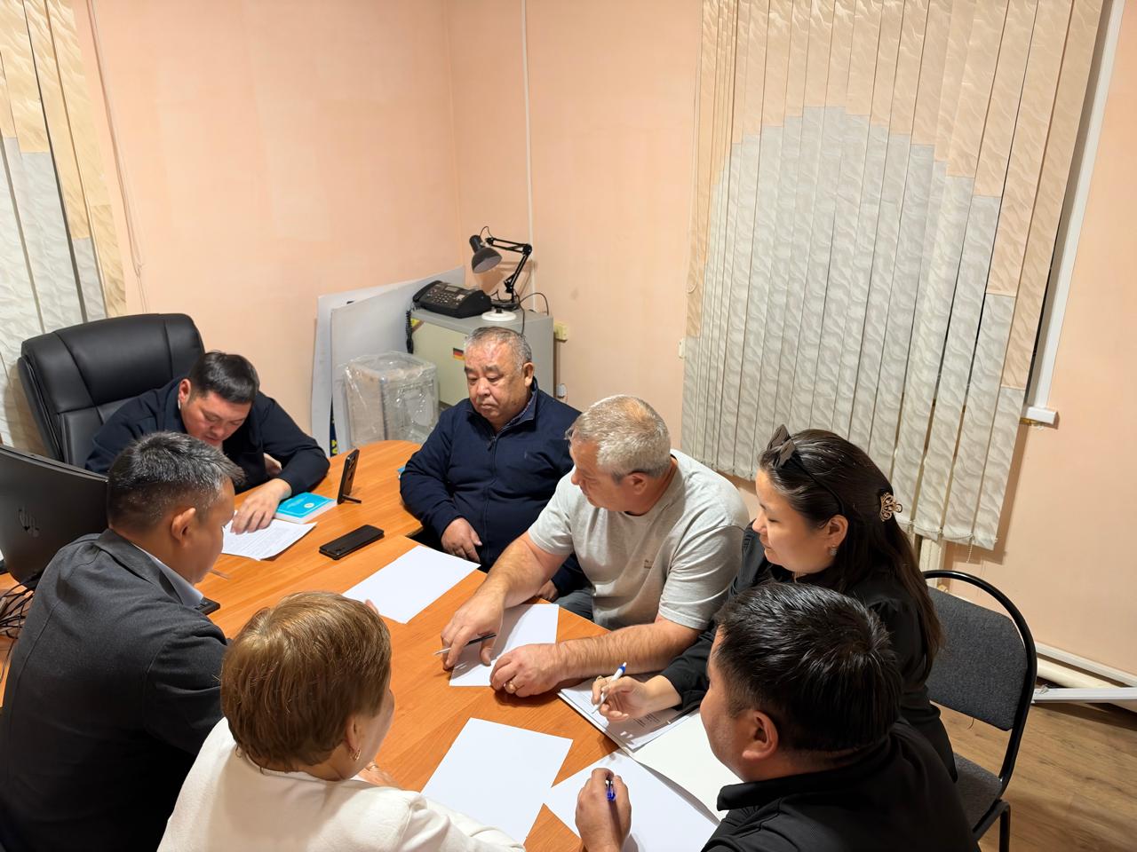 Кеңседе үстел басында жиналып, құжаттармен жұмыс істеп жатқан адамдар тобы.