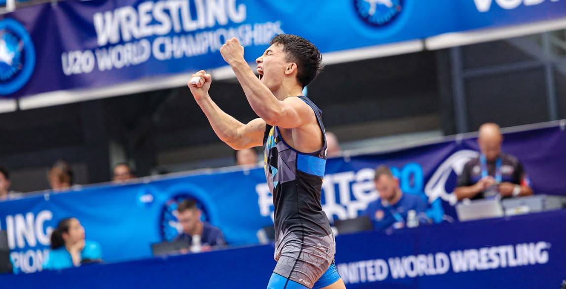 Жеңіске жеткеніне қуанған күресші. Фонда "U20 World Championships" жазуы бар баннер.