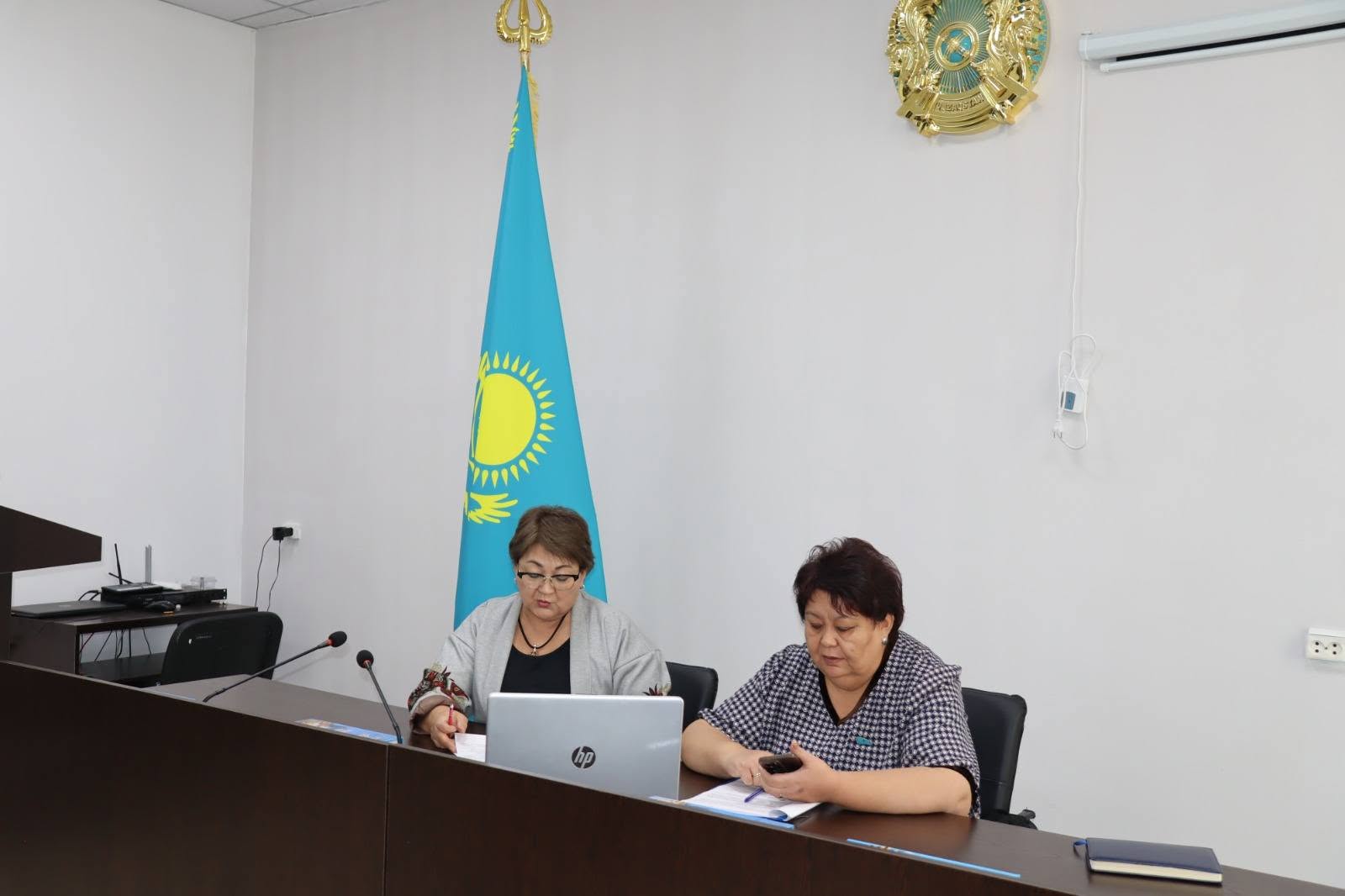Қазақстан туы мен елтаңбасы бар бөлмеде үстел басында отырған екі әйел.