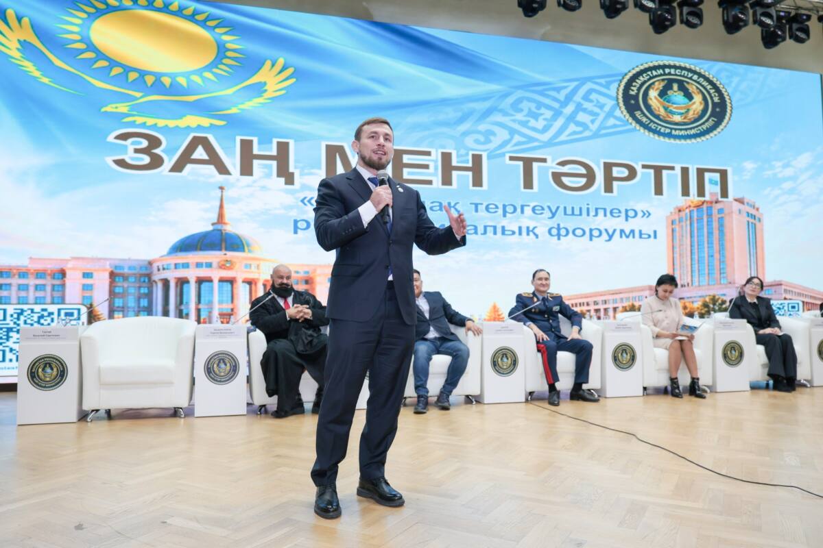Мужчина в костюме выступает с речью на форуме «Заң мен тәртіп» на фоне баннера с государственной символикой Казахстана.