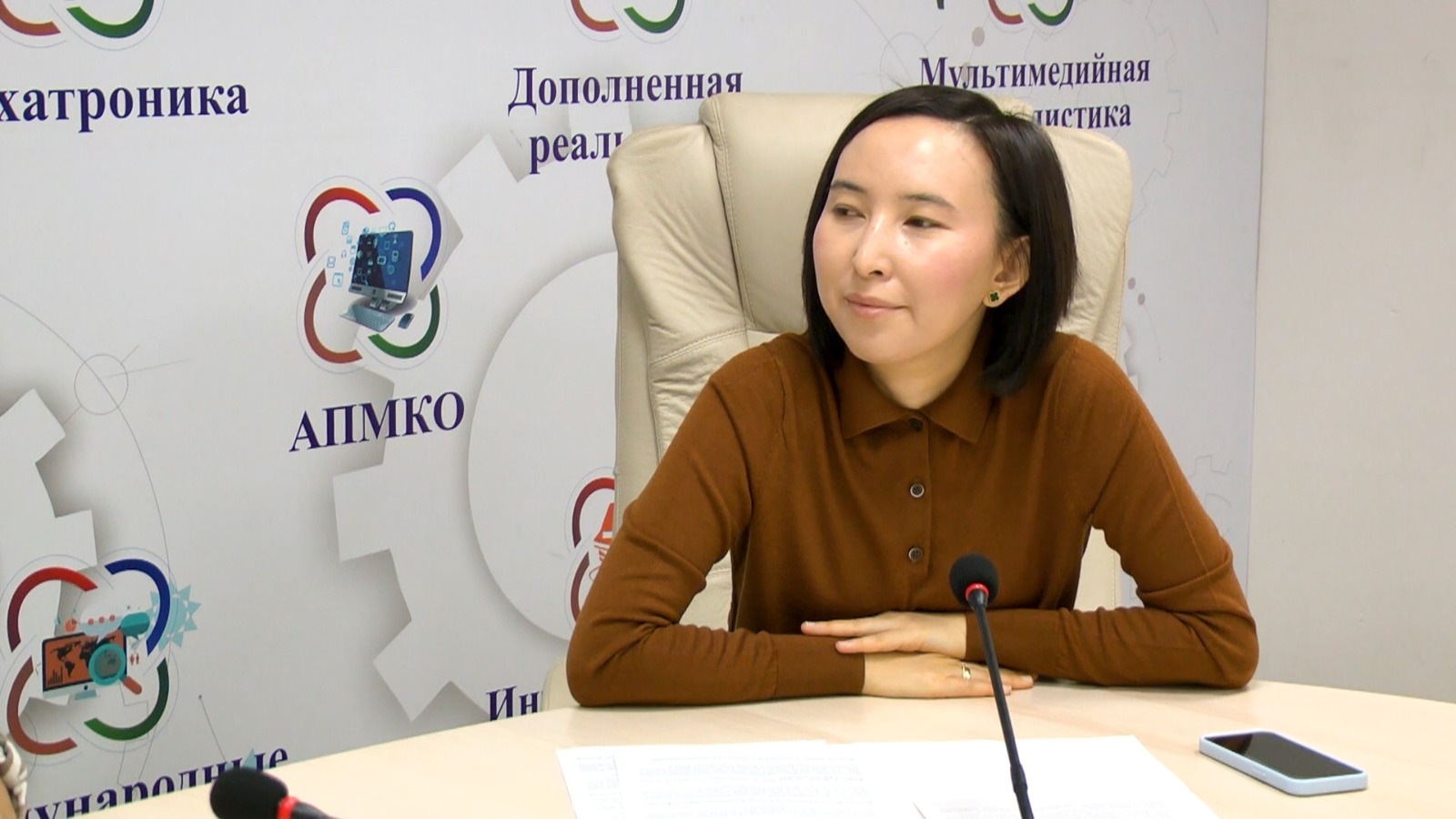 Қоңыр түсті киімдегі әйел адам микрофонның алдында отыр. Артында АПМКО логотипі бар баннер тұр.