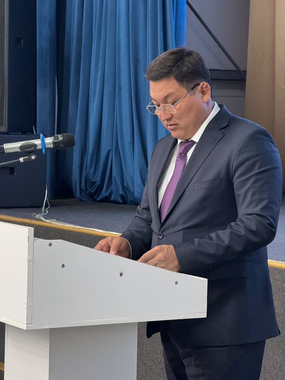 Кілемді және көк перделі фонда, ақ трибуна артында сөйлеп тұрған костюм киген ер адам.