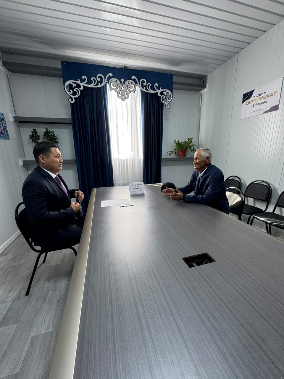 Келіссөз жүргізіп жатқан екі ер адамның кеңседегі суреті.