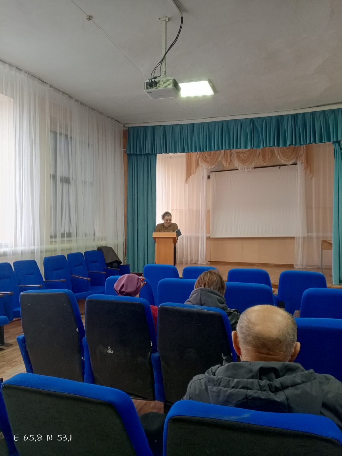 Женщина выступает с речью за трибуной в актовом зале перед аудиторией, сидящей в синих креслах.