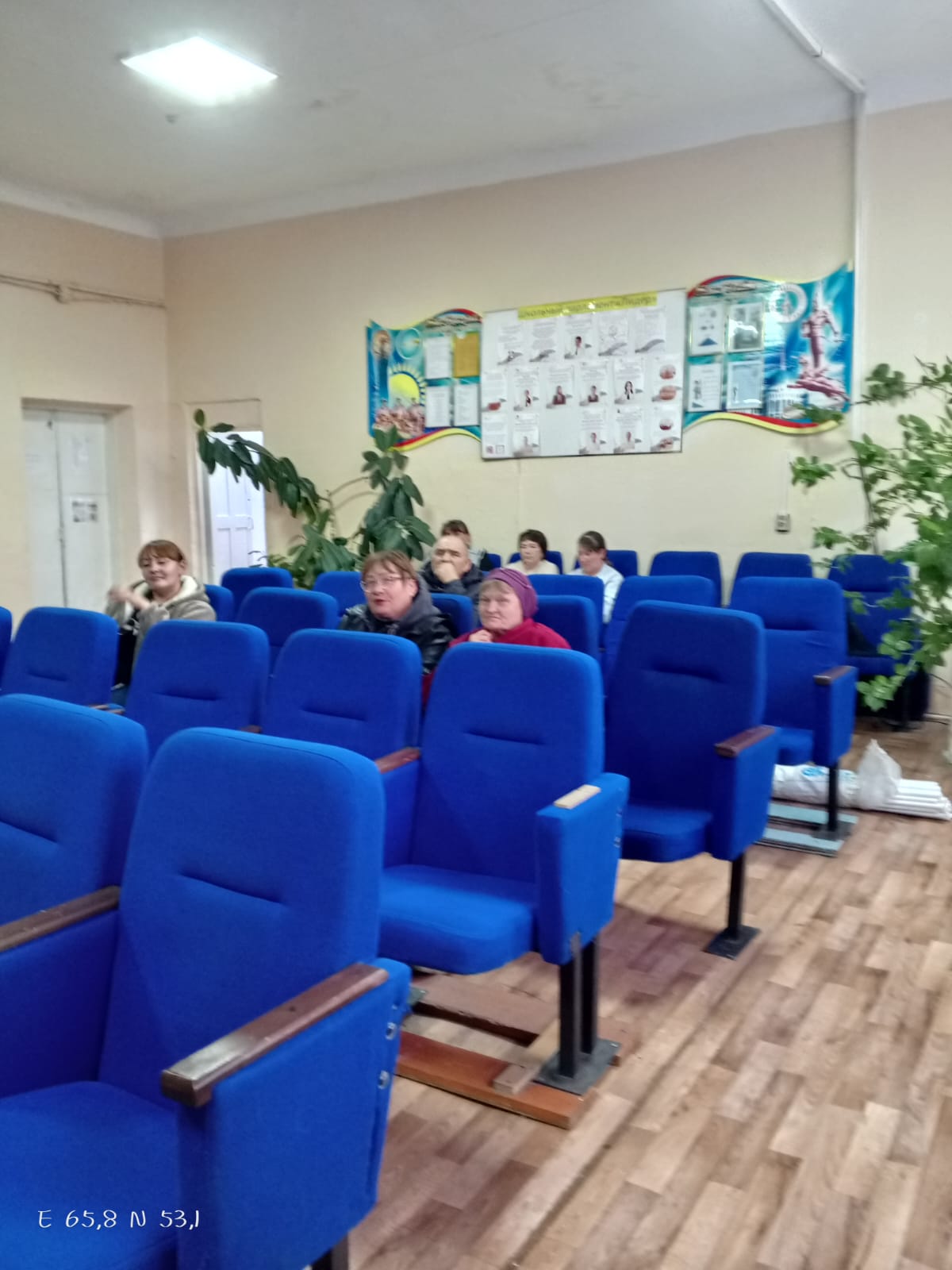 Көк орындықтары бар аудиторияда отырған адамдар.