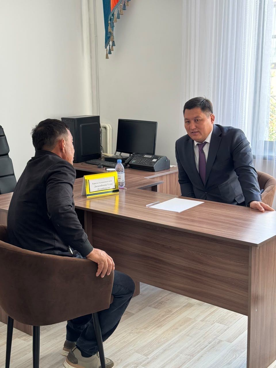 Кеңседе үстел басында бетпе-бет сөйлесіп отырған екі ер адам.