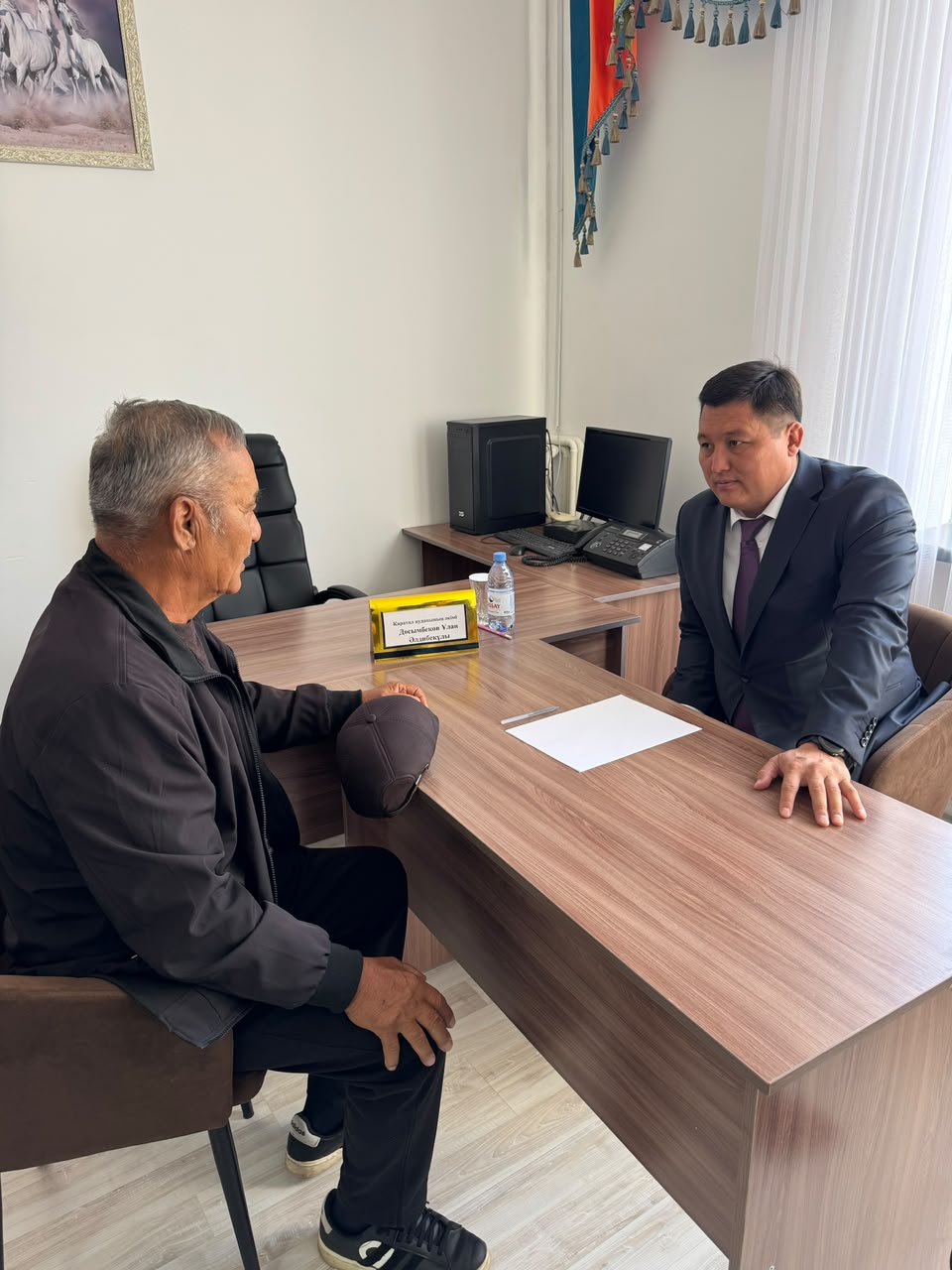 Кеңседе үстел басында бетпе-бет сөйлесіп отырған екі ер адам.