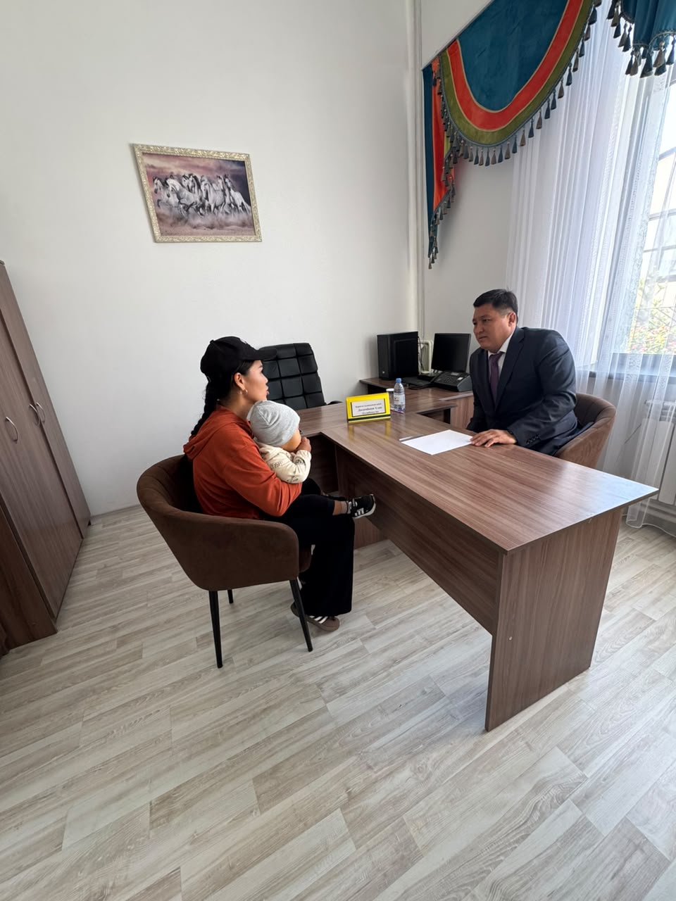 Кеңседе сәбиімен бірге ресми тұлғадан кеңес алып жатқан әйел.