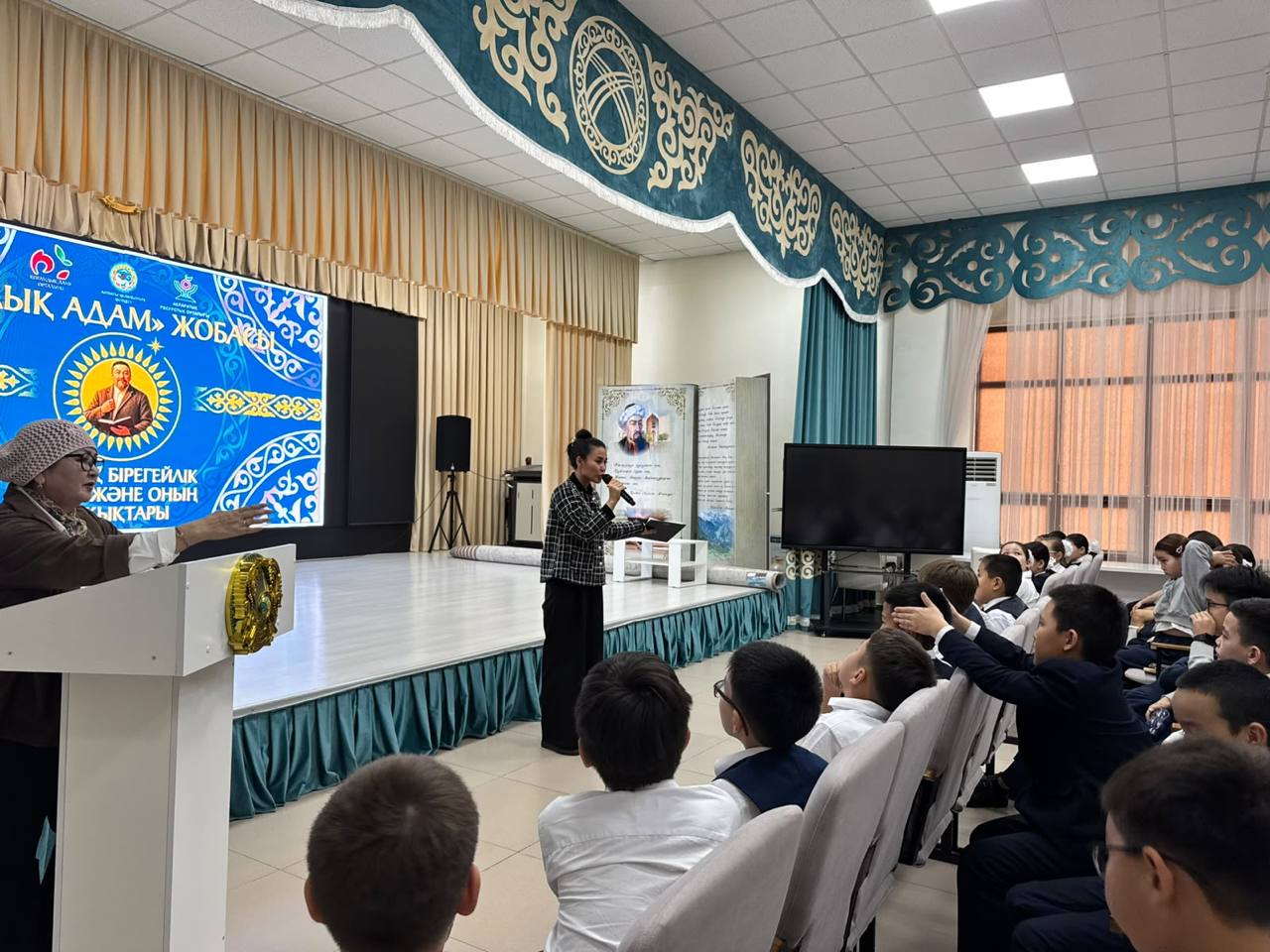 Мектеп акт залындағы іс-шара: сахнада сөйлеп тұрған қыз және оны тыңдап отырған оқушылар.
