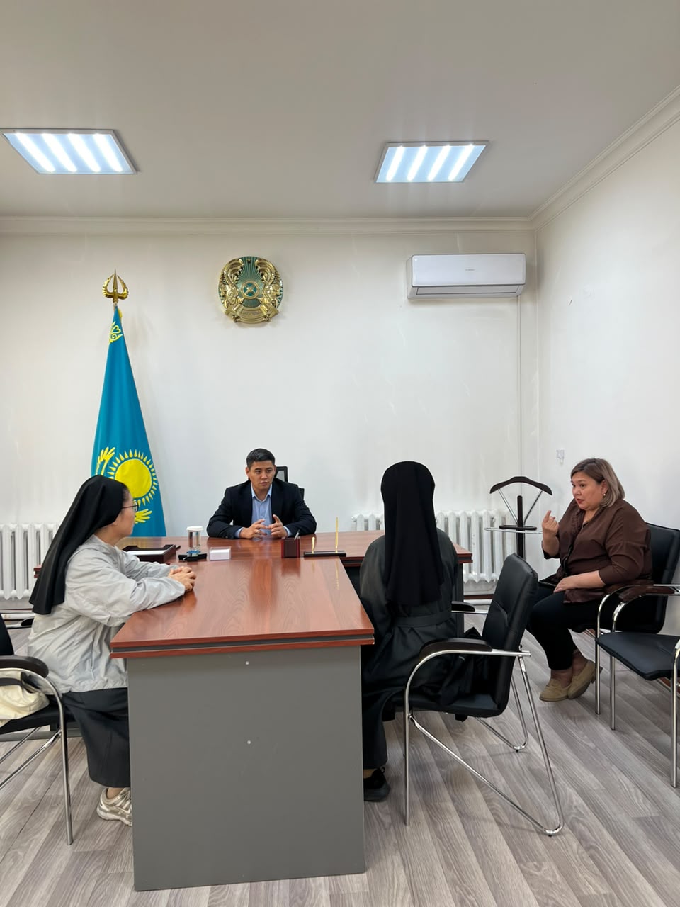 Келіссөз жүргізіп жатқан төрт адам: үстел басындағы ер адам және онымен сөйлесіп отырған үш әйел. Фонда Қазақстан туы мен гербі.
