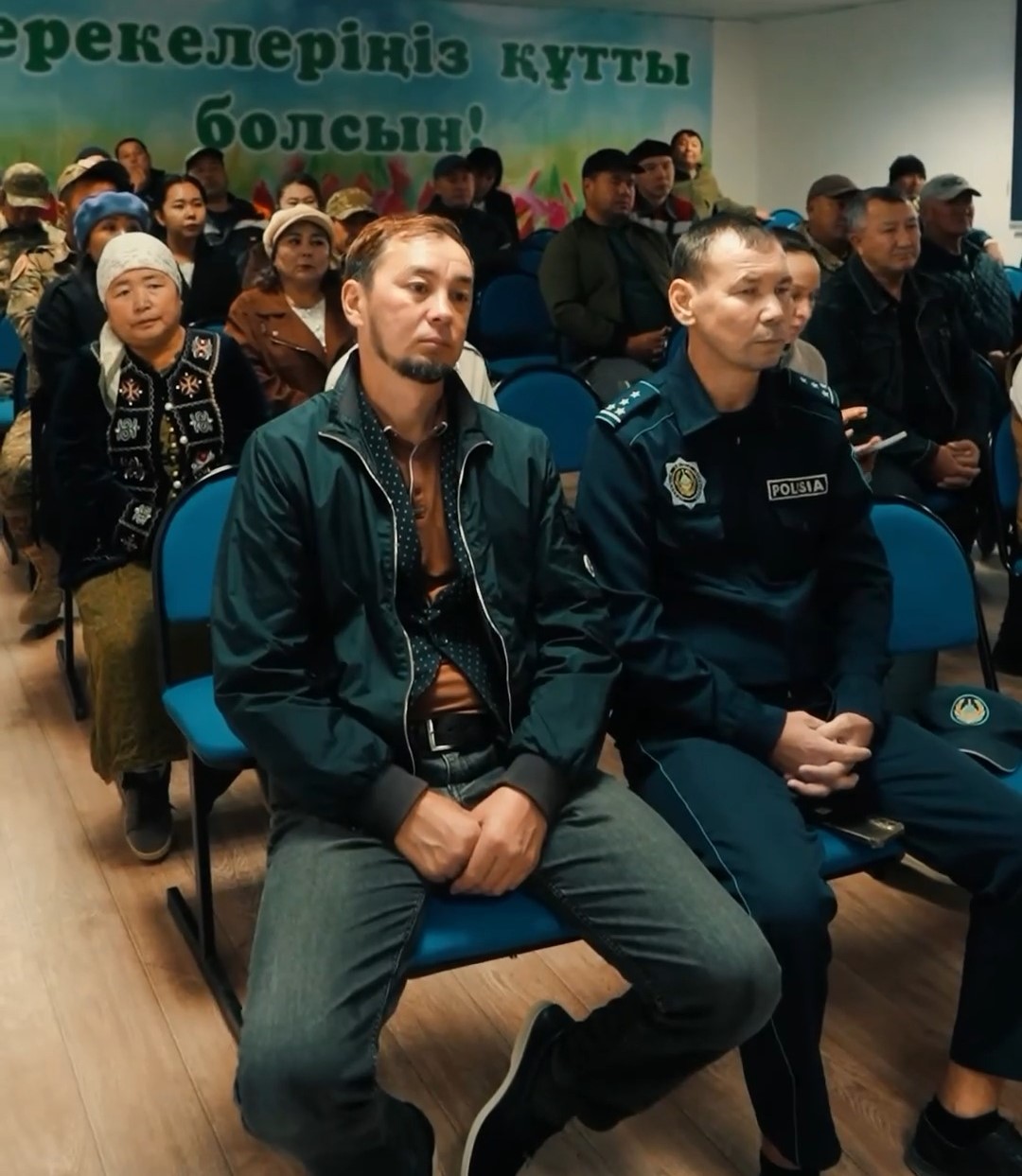 Көк орындықтарда отырған адамдар мен полиция қызметкері.