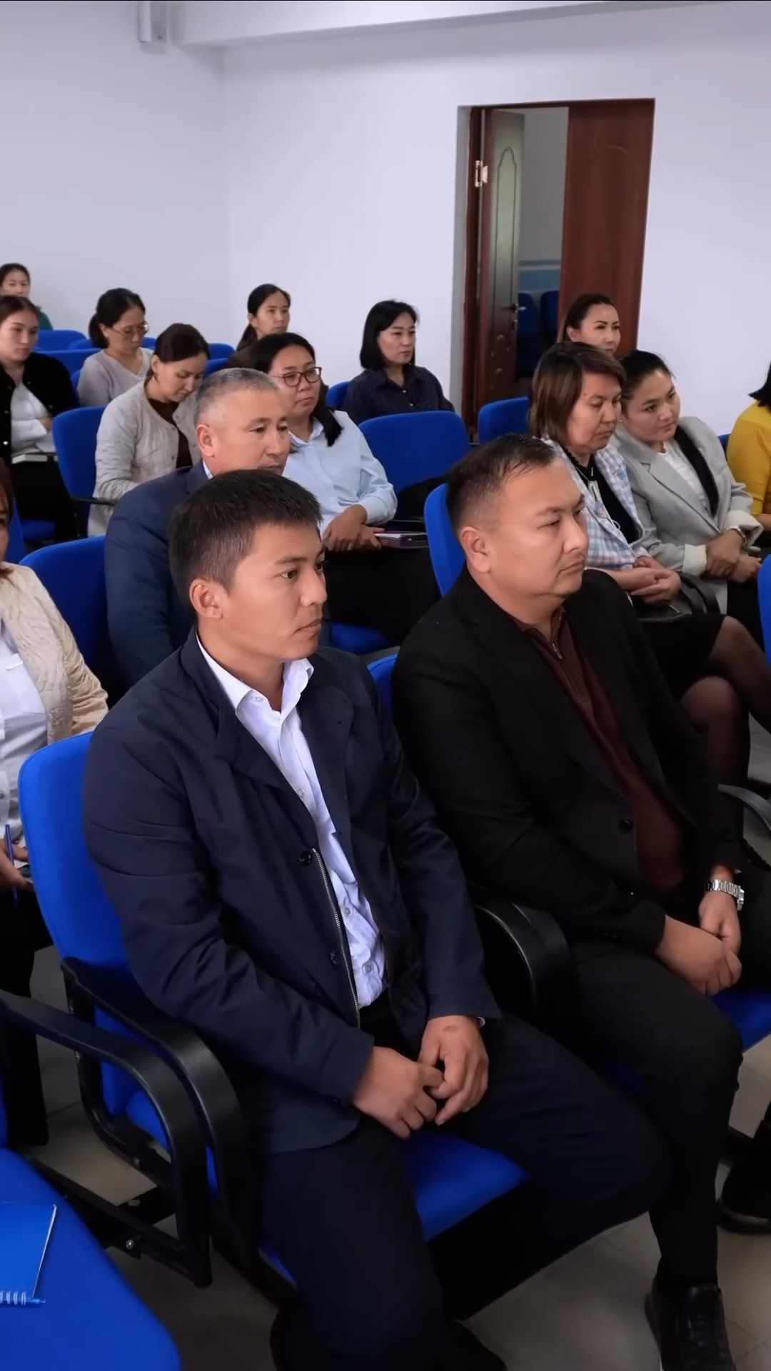 Көк орындықтарда отырған, ресми киінген адамдар тобы.