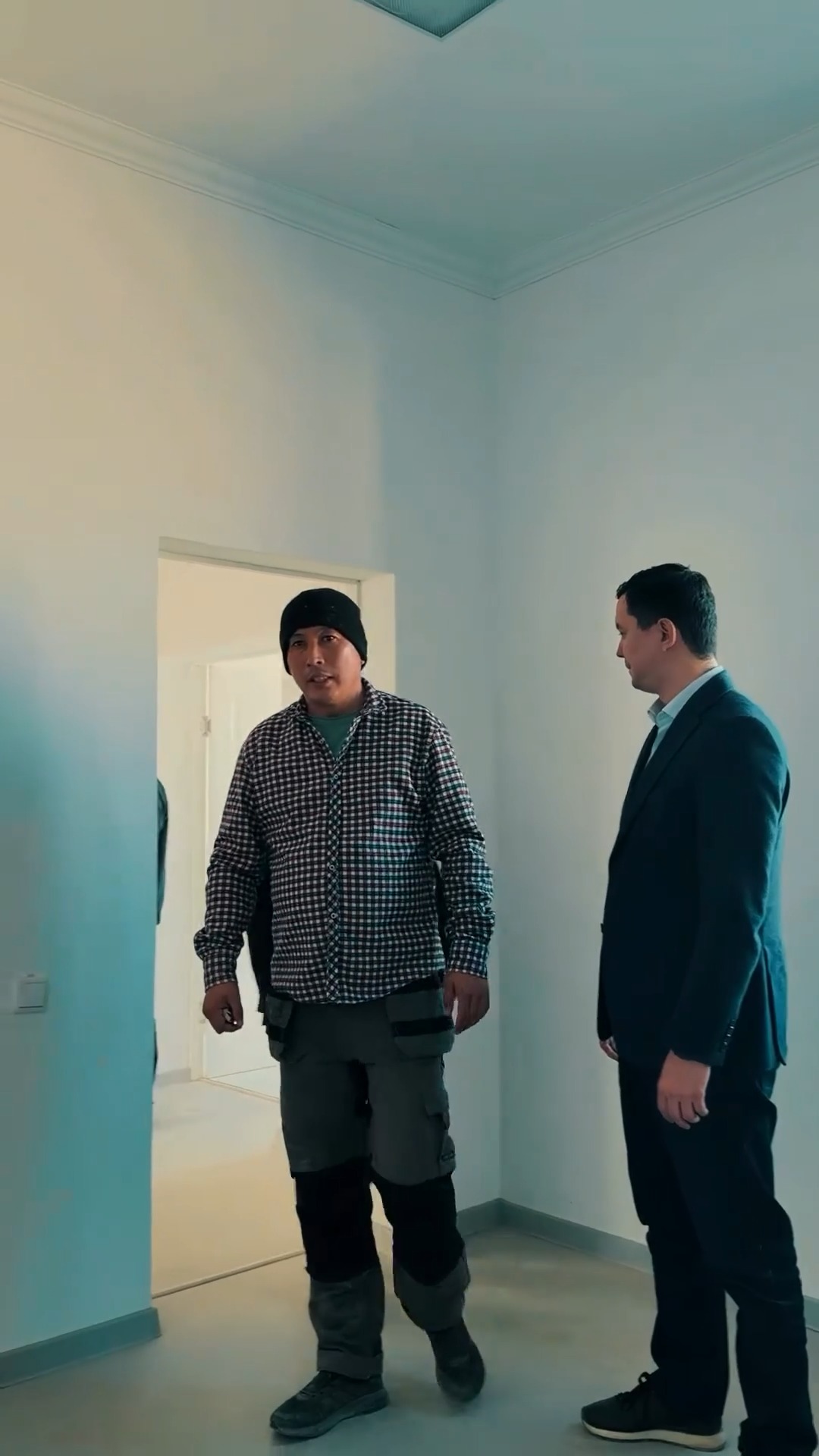 Жұмыс киіміндегі ер адам мен костюм киген ер адамның бөлме ішіндегі сөйлесуі.
