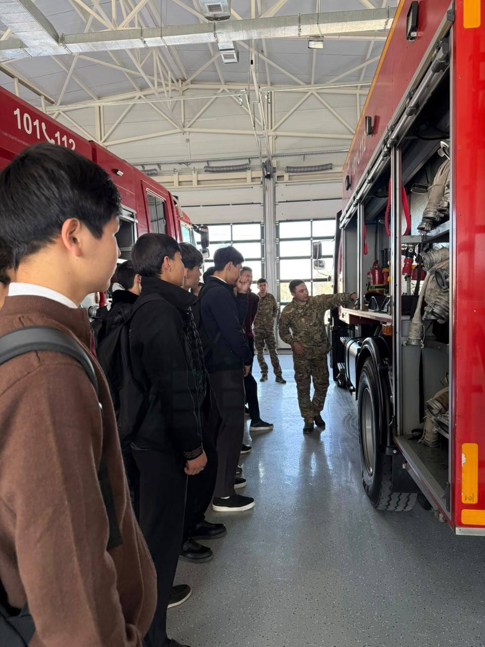 Өрт сөндіру бөлігіндегі өрт сөндіру көлігінің жабдықтарын зерттеп жатқан студенттер тобы және нұсқаушы.