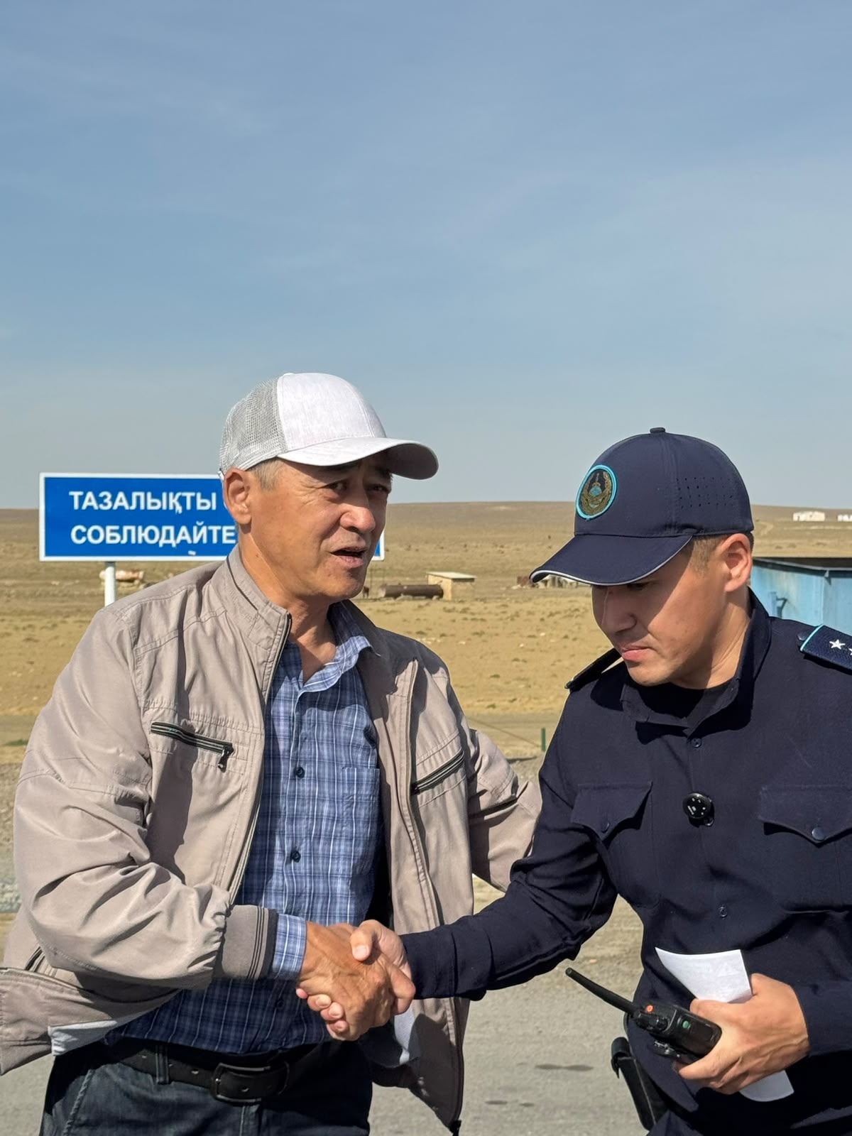 Пожилой мужчина в кепке и полицейский в форме жмут друг другу руки на фоне открытой местности.
