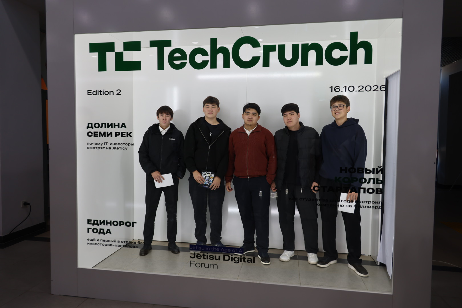 Пятеро молодых людей позируют на фоне баннера TechCrunch Edition 2 на форуме Jetisu Digital Forum.