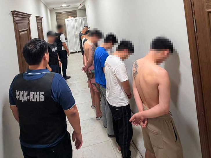 Группа задержанных мужчин стоит лицом к стене в коридоре под присмотром сотрудников в форме.